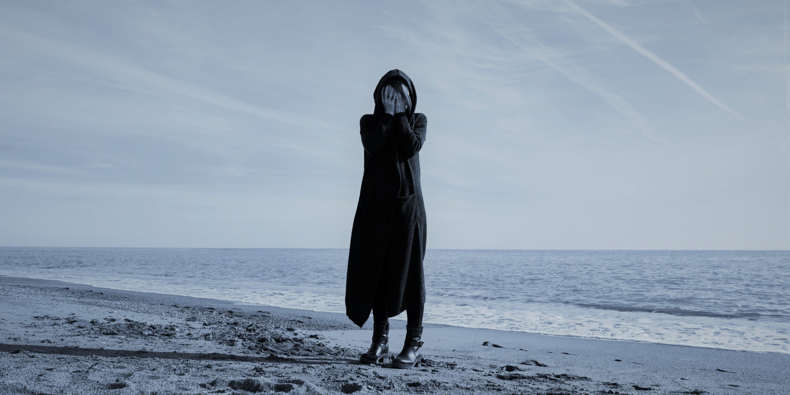 A woman in a dark black hood standing on a beach and covering her face with her hands