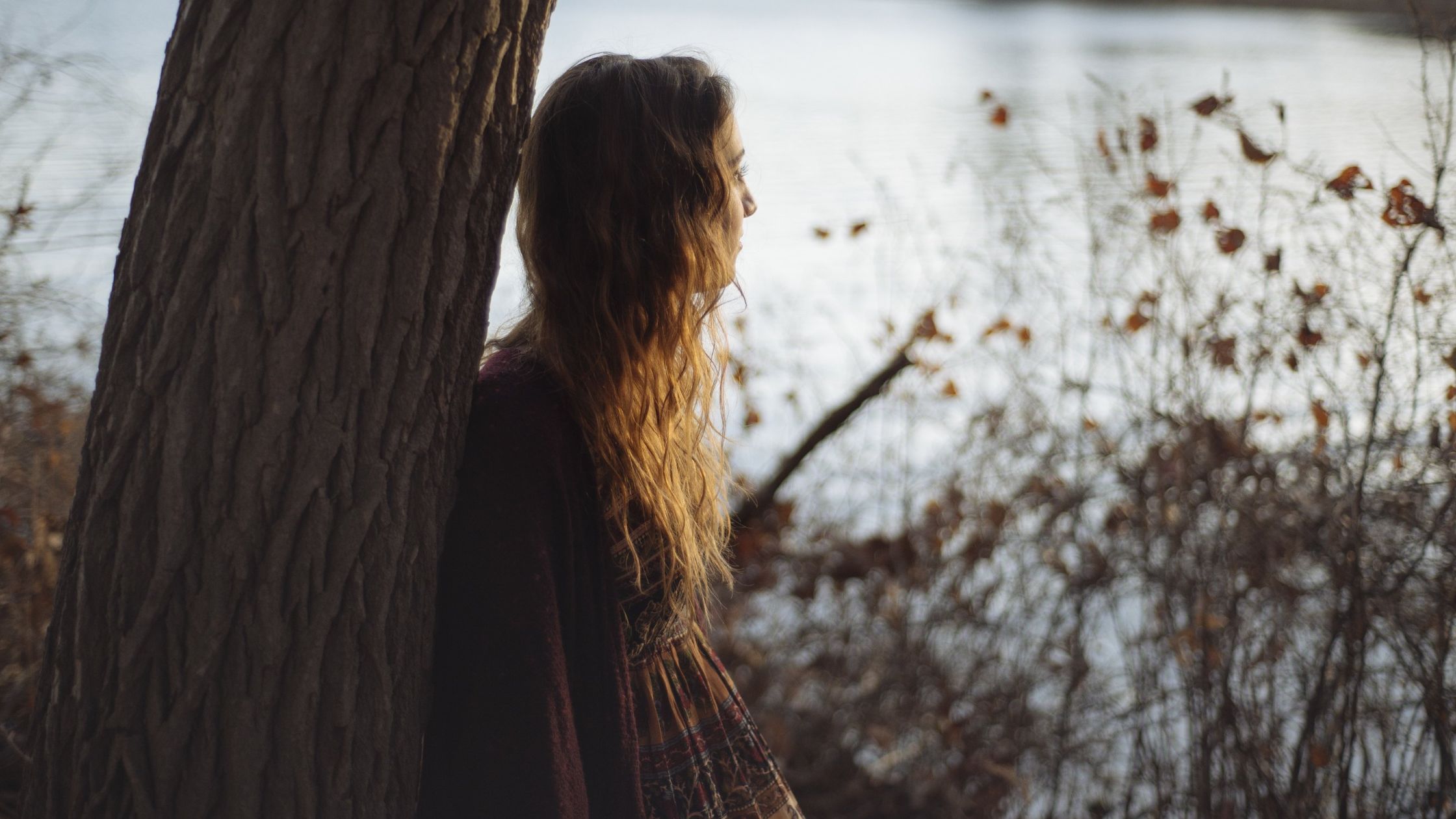 A woman leaning against a tree and staring out a a pond